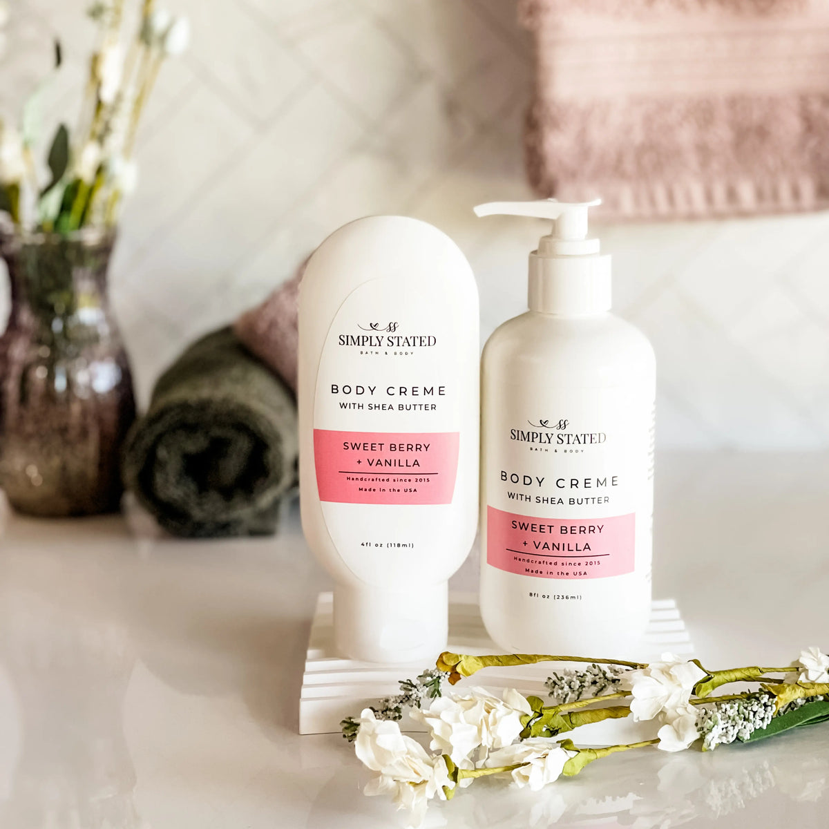 Simply Stated Bath & Body Sweet Berry + Vanilla Body Creme with Shea Butter bottles, styled with towels and flowers