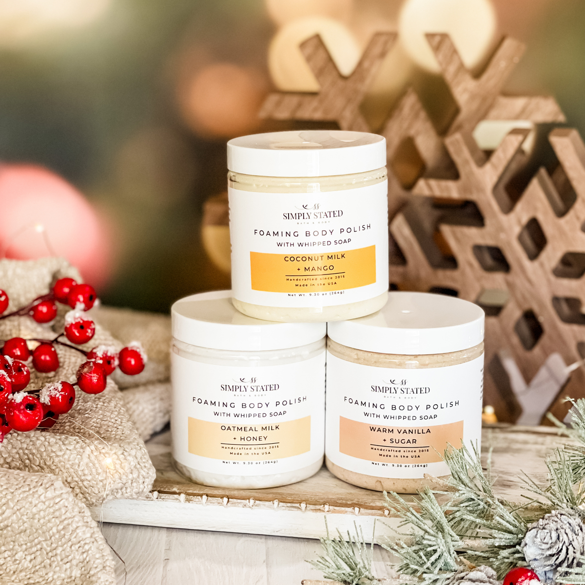 Simply Stated Foaming Body Polish trio with whipped soap, stacked beside wooden snowflakes, red berries, and frosted pine.