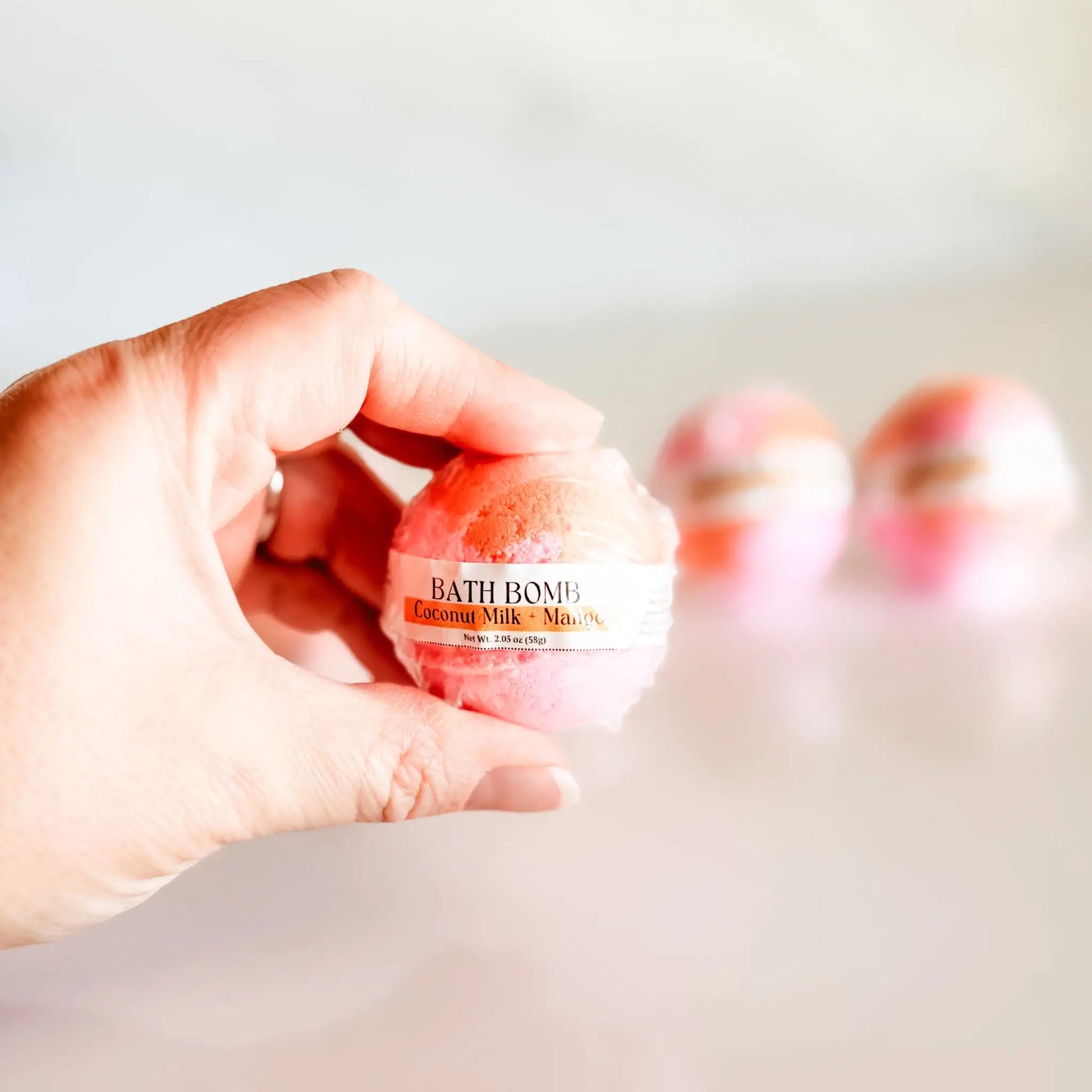 Bath Bomb in Coconut Milk + Mango, pink and orange bath bomb held in hand with more bombs blurred in background