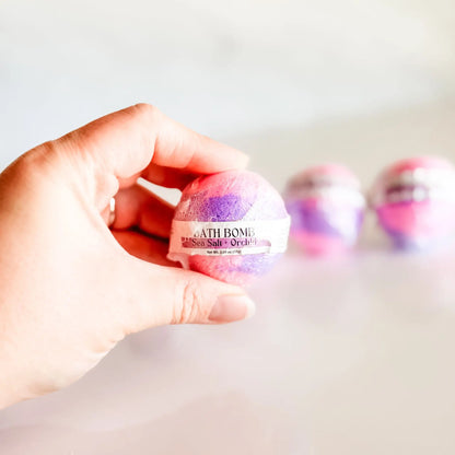 Bath Bomb in Sea Salt + Orchid, pink and purple bath bomb held in hand with more bombs blurred in background