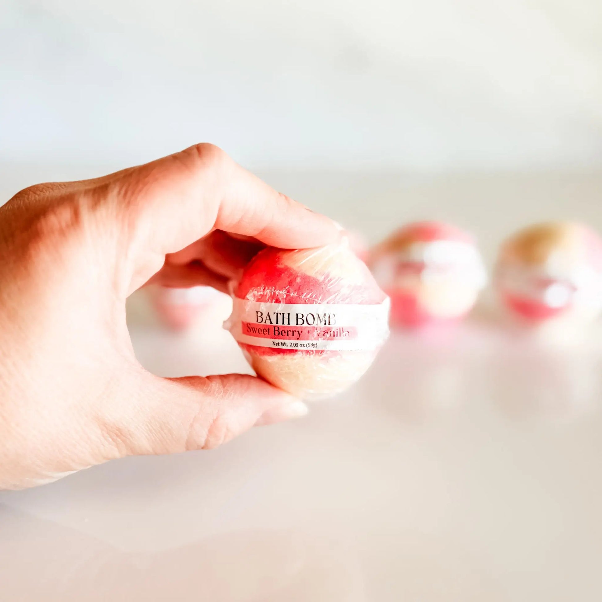Bath Bomb in Sweet Berry + Vanilla, red and white bath bomb held in hand with more bombs blurred in background