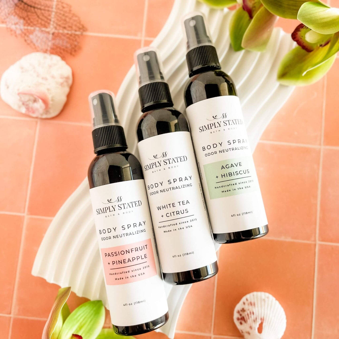 Trio of Simply Stated Summer Collection Body Sprays—Passionfruit + Pineapple, White Tea + Citrus, and Agave + Hibiscus—styled on a white wave tray with tropical decor and shells.