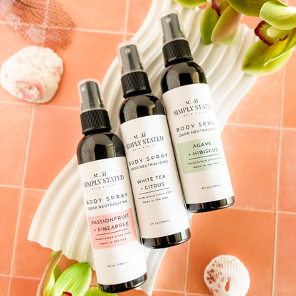 Trio of Simply Stated Summer Collection Body Sprays—Passionfruit + Pineapple, White Tea + Citrus, and Agave + Hibiscus—styled on a white wave tray with tropical decor and shells.