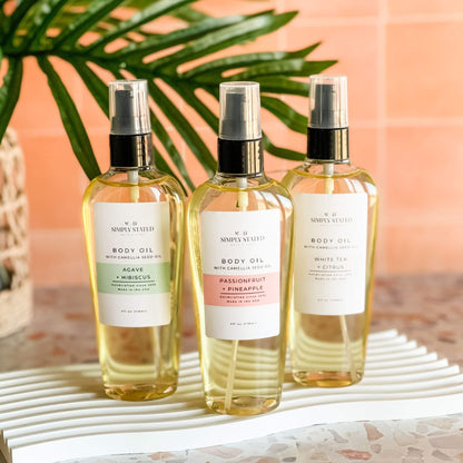 Trio of Simply Stated Bath & Body oils in Agave + Hibiscus, Passionfruit + Pineapple, and White Tea + Citrus. Styled on a white tray with a tropical backdrop. Camellia seed oil body moisturizers with summery scent profiles and clean branding.