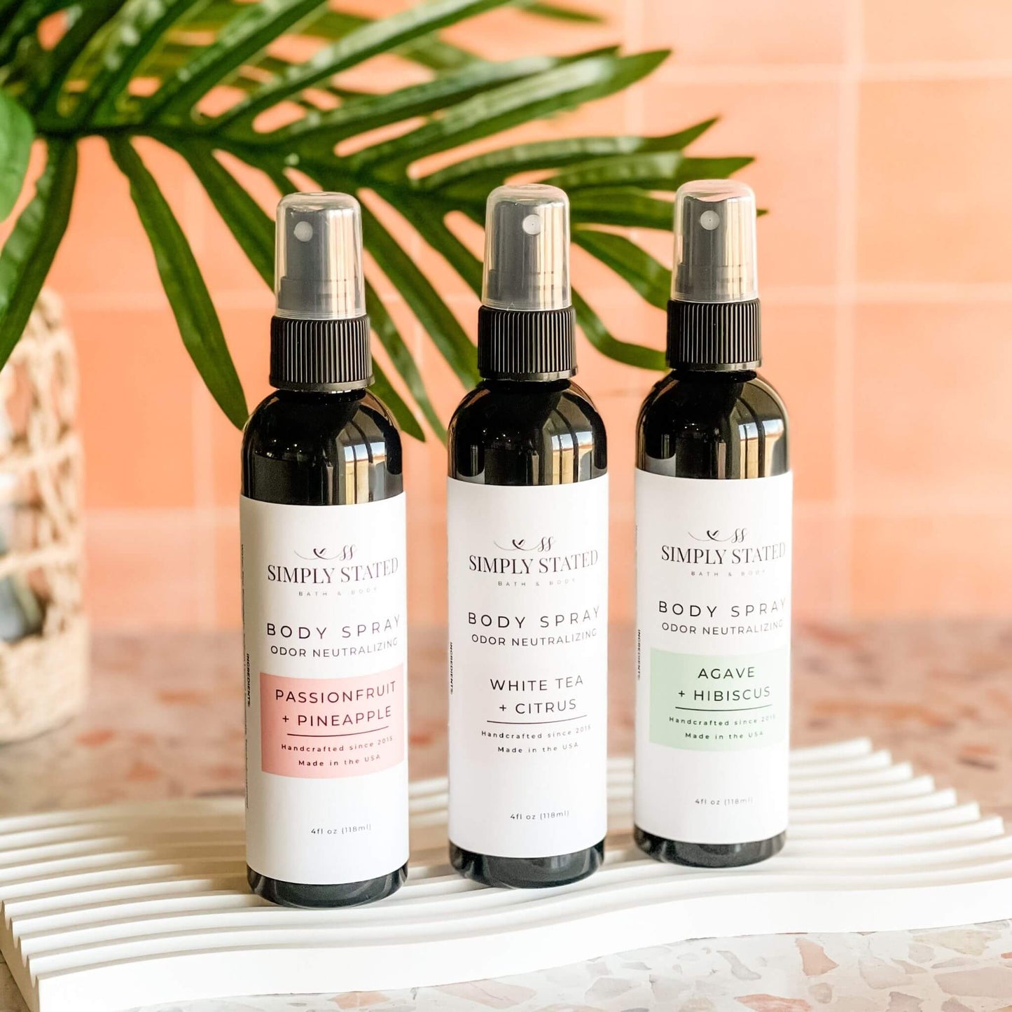 Simply Stated Summer Body Spray trio displayed upright—Passionfruit + Pineapple, White Tea + Citrus, and Agave + Hibiscus—against a tropical backdrop with palm leaf decor.
