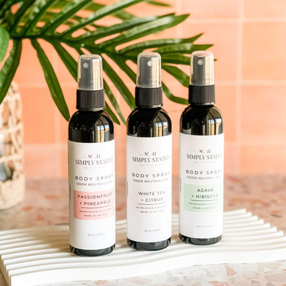 Simply Stated Summer Body Spray trio displayed upright—Passionfruit + Pineapple, White Tea + Citrus, and Agave + Hibiscus—against a tropical backdrop with palm leaf decor.