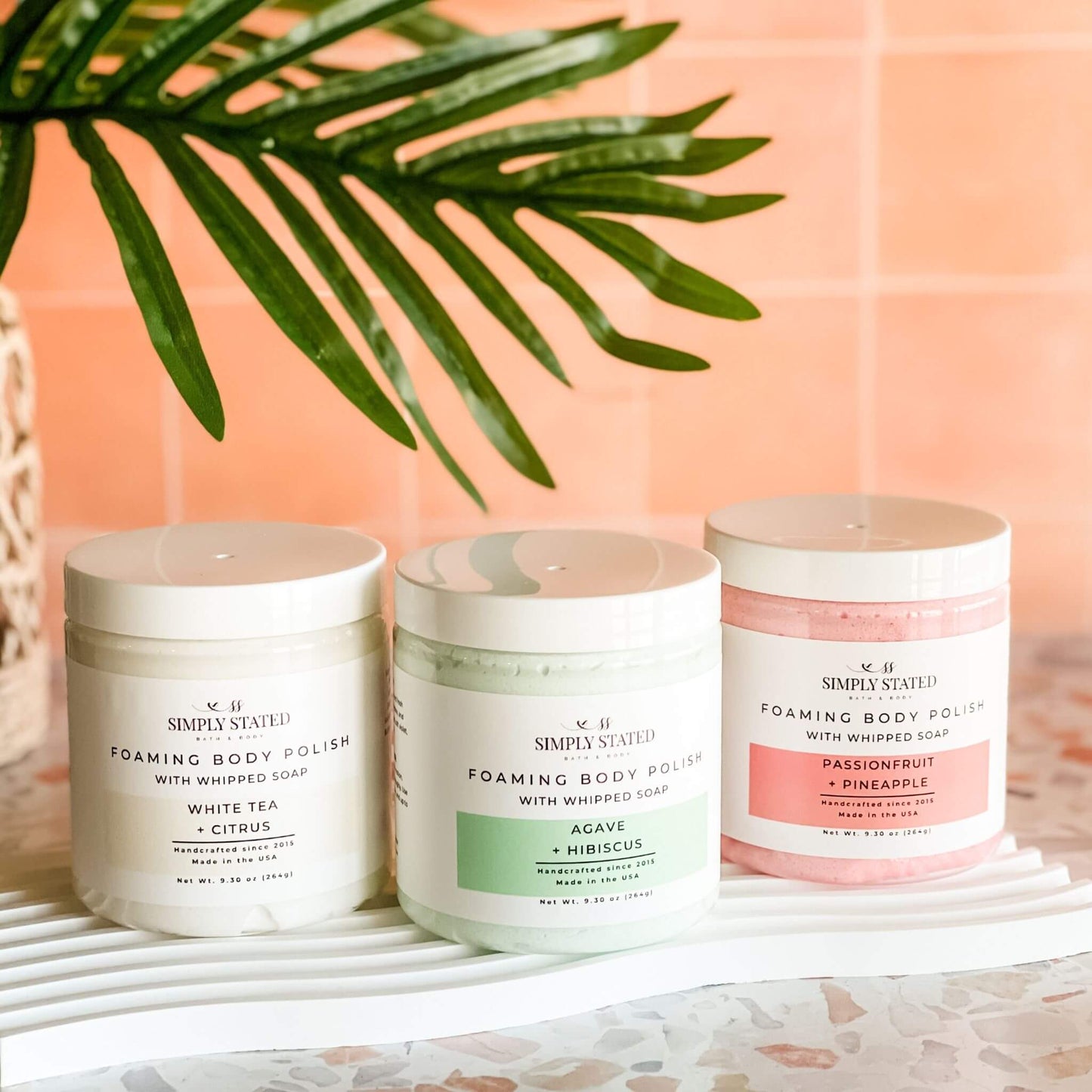 Summer collection of Simply Stated Foaming Body Polishes – three 9.30 oz jars in Passionfruit + Pineapple, Agave + Hibiscus, and White Tea + Citrus, styled with tropical greenery.