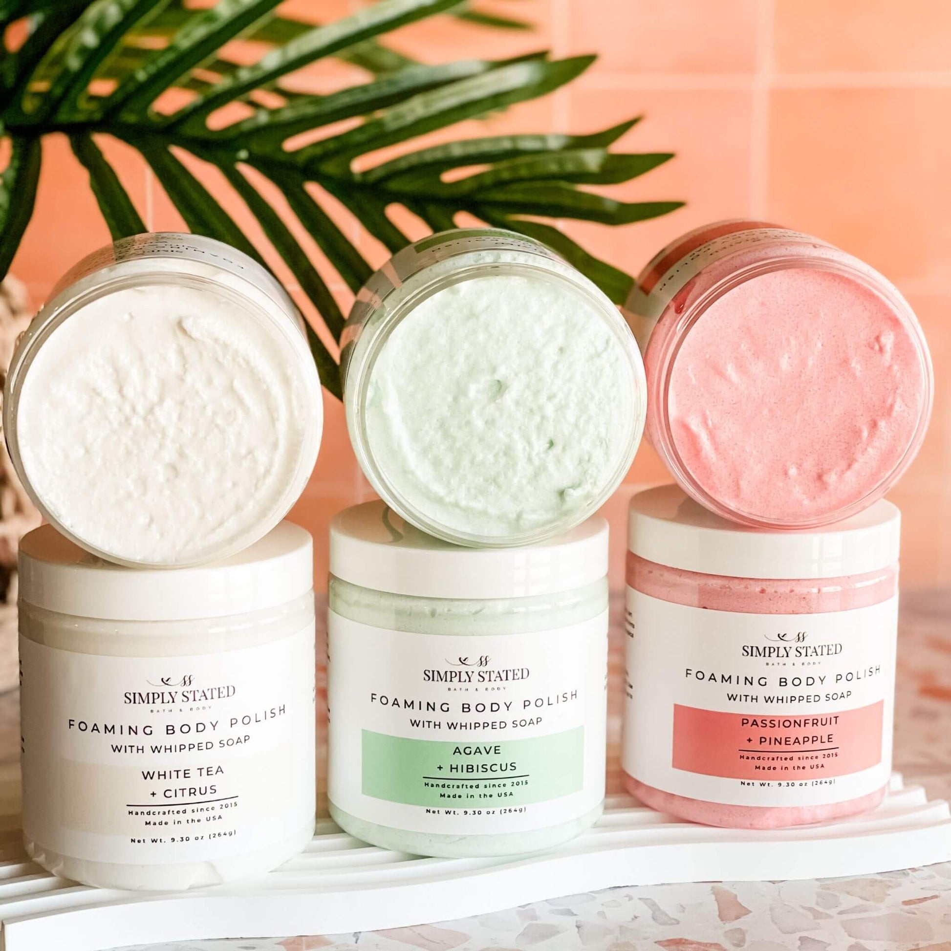 Trio of Simply Stated Foaming Body Polishes with open jars displaying whipped textures – tropical Passionfruit + Pineapple, fresh Agave + Hibiscus, and citrusy White Tea + Citrus, styled on a terrazzo surface.