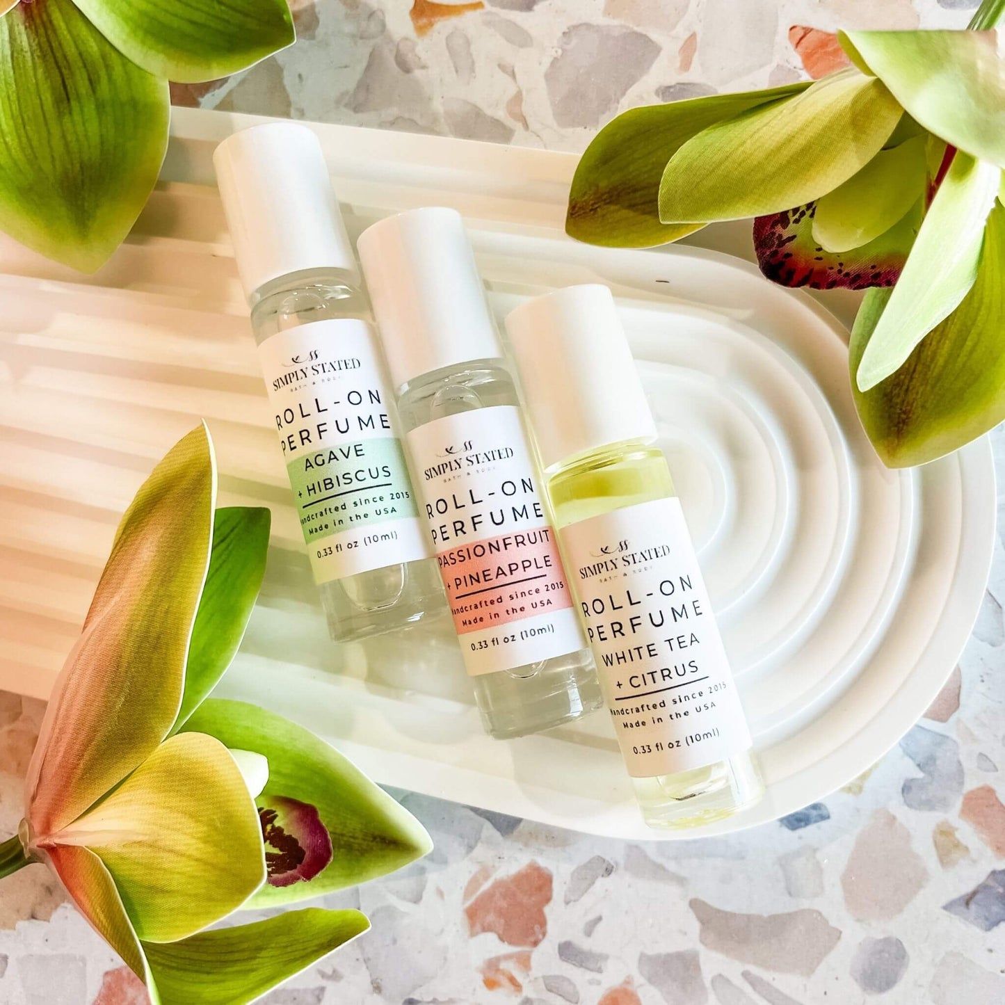 Simply Stated Summer Collection Roll-On Perfumes in Agave + Hibiscus, Passionfruit + Pineapple, and White Tea + Citrus – flat lay display with orchid flowers and terrazzo surface.