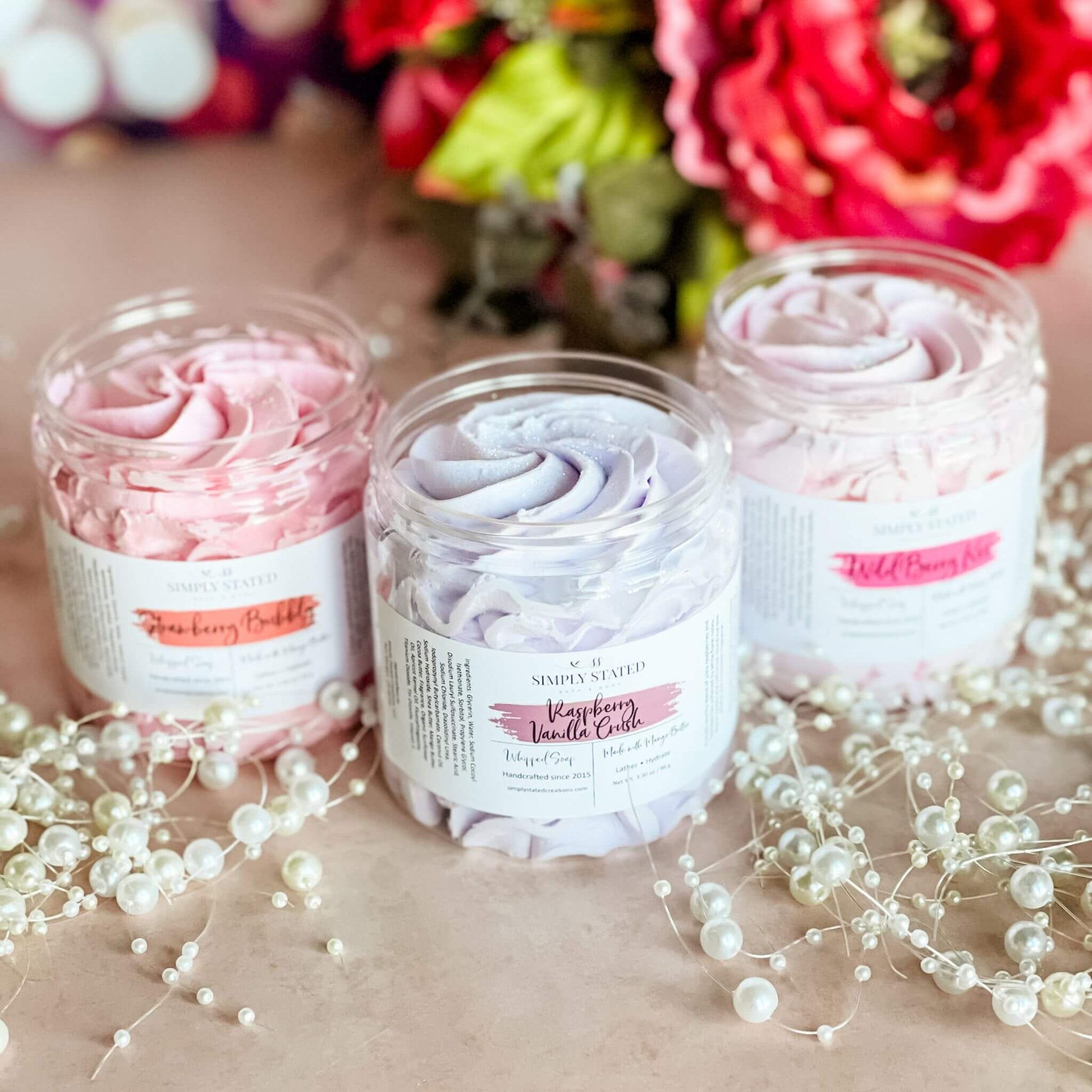 A trio of Sweet Temptations Whipped Soaps—Raspberry Vanilla Crush, Strawberry Bubbly, and Wild Berry Kiss—arranged with pearl accents and vibrant floral decor on a blush pink background.