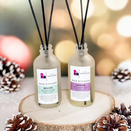 The Block x Simply Stated Reed Diffusers for rooms featuring scents in Revive and Gather with a warm winter glow background and pinecone decorations.