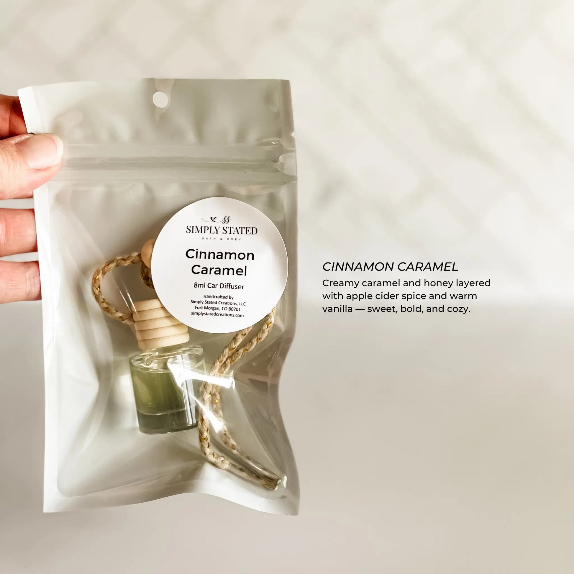 Hand holding a 'Simply Stated' cinnamon caramel car air freshener package on a light background