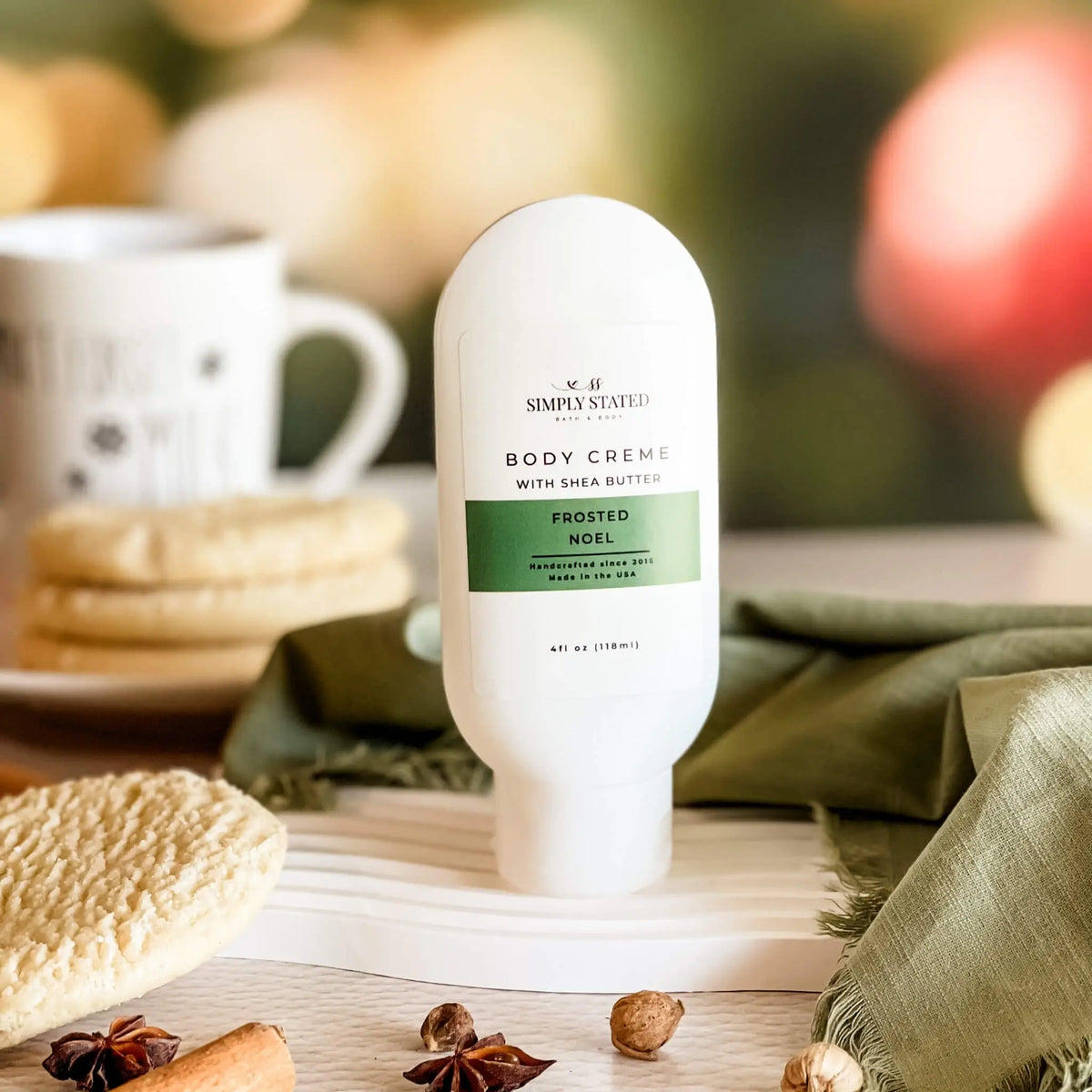 Simply Stated Frosted Noel Body Creme with shea butter, cozy holiday scene with cinnamon, star anise, and mug.