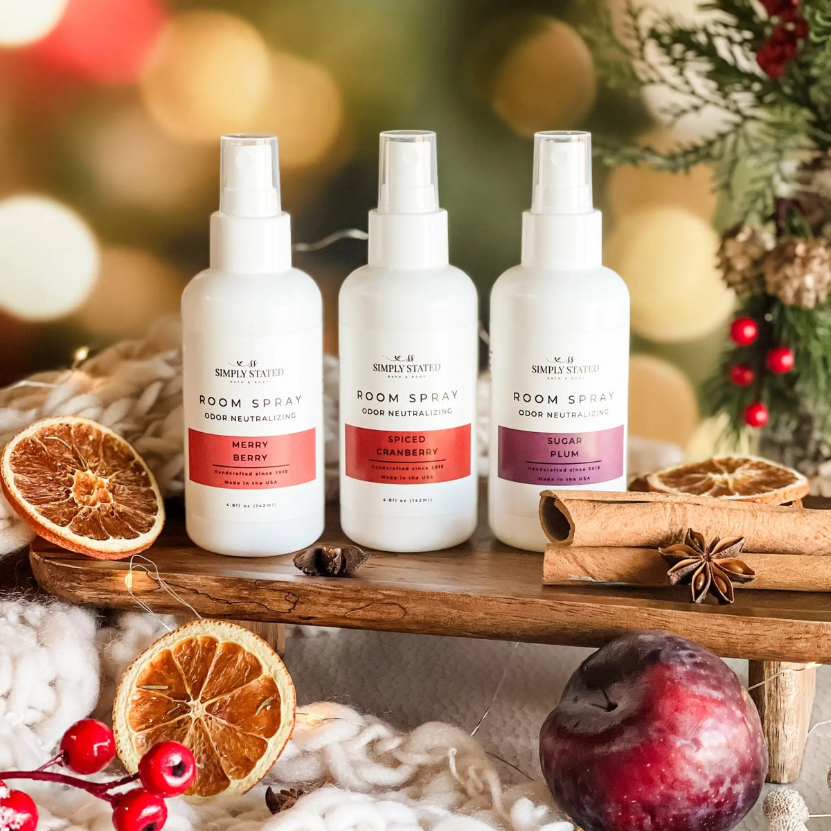 Simply Stated Room Spray trio, Merry Berry, Spiced Cranberry, Sugar Plum, on a wood shelf with cinnamon and plum.