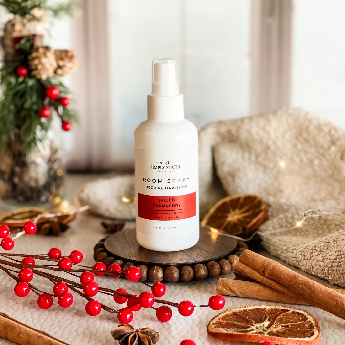 Room spray bottle on a wooden coaster with festive decorations including red berries and cinnamon sticks.